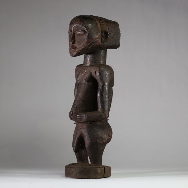 Statue Hemba Singiti, RD Congo. La statue représente une figure masculine debout, dans une posture frontale hiératique. La tête, surdimensionnée, est marquée par une barbe trapézoïdale finement striée, un visage calme aux yeux mi-clos, un nez droit et une bouche discrète. À l’arrière du crâne, une coiffe cruciforme profondément sculptée suggère une coiffure cérémonielle typique. Les bras sont plié