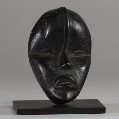 Masque passeport Dan, Côte d’Ivoire. Bois. Il présente une esthétique d’une grande pureté. Le visage, de forme ovale, est sculpté avec sobriété : les yeux sont mi-clos et incisés en amande, le nez est droit, triangulaire et structuré, et la bouche légèrement ouverte laisse deviner une expression intériorisée. Une ligne verticale en relief s’élève du haut du nez jusqu’au front, accentuant l’axe de