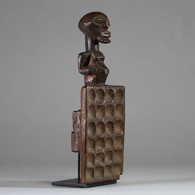 Plateau de jeu type « awalé » rituel Songye, RD Congo. Bois ancien, figure anthropomorphe stylisée, dont la partie inférieure est un plateau creusé de 24 alvéoles disposées en quatre rangées de six. Le personnage reprend les canons classiques de la culture Songye avec les bras collés au corps, et un ventre proéminent. Bel objet initiatique et symbolique à la croisée du ludique et du sacral, associ