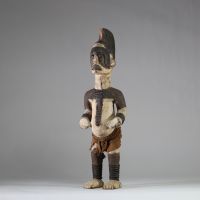 Statue Igbo « Alusi ». Nigéria. Bois, textile. Représentation masculine de type Alusi, divinité secondaire ou esprit tutélaire propre au peuple Igbo (sud-est du Nigeria), vénérée dans des sanctuaires collectifs ou familiaux. Grande crête sagittale sculptée d’une coiffe en éventail semi circulaire, ornée de motifs en spirales. Front bombé, yeux globuleux en amande, sans pupille, sourcils et joues m