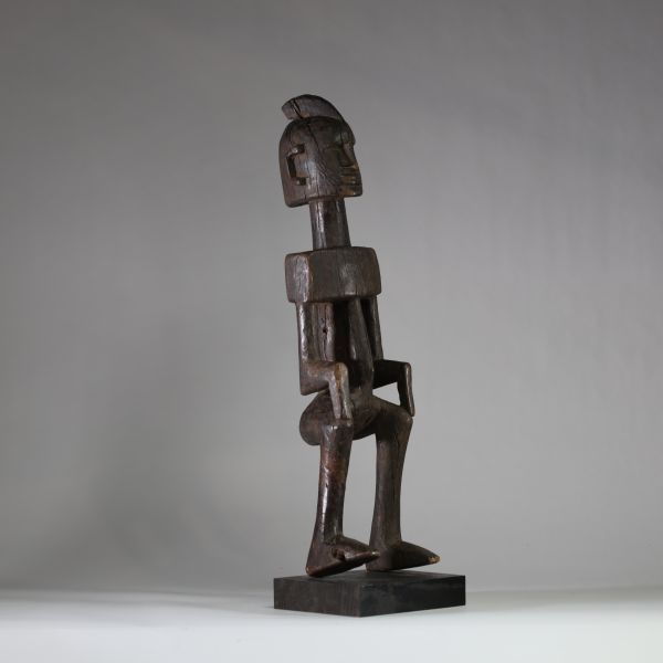 Statue masculine Dogon, Mali . Bois dense. Le style général est typique des productions Dogon anciennes, en particulier celles issues du plateau de Bandiagara. Le traitement géométrique du torse, l’élongation du cou, la posture figée et l’expression sobre indiquent une représentation d’ancêtre ou de figure spirituelle. La coiffe sagittale évoque les attributs cérémoniels du Hogon. La patine est an