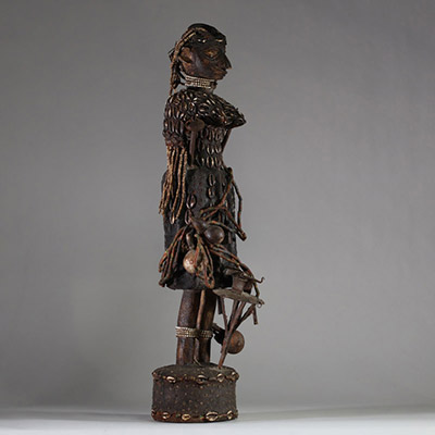 Intéressante statue féminine de type Bocio, Fon, Bénin. Le visage est sculpté de manière stylisée avec un nez proéminent, des yeux en amande profondément incisés, et des lèvres pleines. Une coiffe structurée orne la tête, enrichie de cauris incrustés et de perles. Le cou porte plusieurs rangs de colliers perlés. Le torse et le haut du corps sont entièrement recouverts de cauris patinés, suggérant