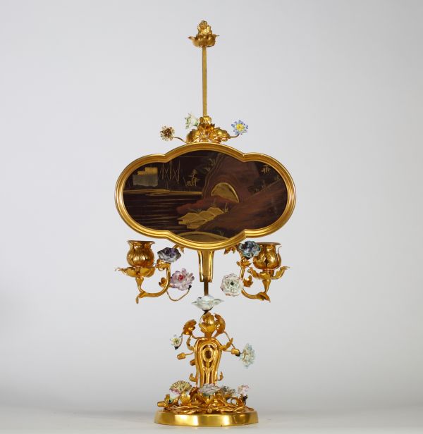 Bougeoir à écran, en bronze ciselé et doré, à décor ajouré de rinceaux feuillagés ponctués de fleurs en porcelaine polychrome de style Meissen. L’ensemble est surmonté d’un écran en laque du Japon de forme chantournée, à décor de paysage lacustre doré dans le goût des exportations de l’époque Meiji. Travail pouvant être attribué à la prestigieuse maison parisienne de l’Escalier de Cristal, vers 18