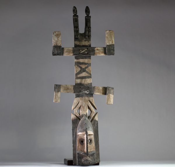 Grand masque Dogon « Kanaga », Mali. Bois ancien et sec, avec des pigments naturels noirs et blancs appliqués de manière traditionnelle. Le visage stylisé présente des yeux en triangle évidés, surmonté d'une superstructure composée de deux barres transversales et de deux personnages anthropomorphes sculptés au sommet. La structure générale respecte les canons de la statuaire Dogon liée aux cérémon
