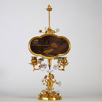 Bougeoir à écran, en bronze ciselé et doré, à décor ajouré de rinceaux feuillagés ponctués de fleurs en porcelaine polychrome de style Meissen. L’ensemble est surmonté d’un écran en laque du Japon de forme chantournée, à décor de paysage lacustre doré dans le goût des exportations de l’époque Meiji. Travail pouvant être attribué à la prestigieuse maison parisienne de l’Escalier de Cristal, vers 18