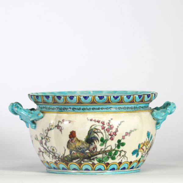 Théodore Deck (1823–1891) Importante jardinière en faïence émaillée, à décor polychrome d’inspiration extrême-orientale représentant un coq, des oiseaux, des papillons et des branchages fleuris en léger relief, sur fond crème. Frise supérieure ornée de motifs géométriques bleus et or. Ansés latérales en forme de dragons stylisés émaillés turquoise.
