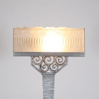 France, vers 1930 Lampe de bureau Art Déco en métal martelé nickelé et verre pressé à décor floral et géométrique.  Reposant sur une base rectangulaire, cette lampe présente un fût élancé à surface martelée, d’une sobriété élégante, souligné d’une finition nickelée mate. L’ensemble est rehaussé d’un nœud ornemental de volutes forgées à la main, typique du vocabulaire décoratif de l’époque.  L’abat