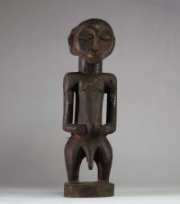 Statue Hemba Singiti, RD Congo. La statue représente une figure masculine debout, dans une posture frontale hiératique. La tête, surdimensionnée, est marquée par une barbe trapézoïdale finement striée, un visage calme aux yeux mi-clos, un nez droit et une bouche discrète. À l’arrière du crâne, une coiffe cruciforme profondément sculptée suggère une coiffure cérémonielle typique. Les bras sont plié