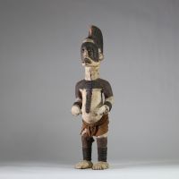 Statue Igbo « Alusi ». Nigéria. Bois, textile. Représentation masculine de type Alusi, divinité secondaire ou esprit tutélaire propre au peuple Igbo (sud-est du Nigeria), vénérée dans des sanctuaires collectifs ou familiaux. Grande crête sagittale sculptée d’une coiffe en éventail semi circulaire, ornée de motifs en spirales. Front bombé, yeux globuleux en amande, sans pupille, sourcils et joues m