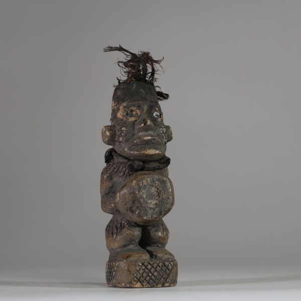 Ancienne figure rituelle en pierre  Bakongo, RD Congo. Travail de sculpture très fin. Le personnage a les mains posées sur le bilogo, protégé par un morceau de miroir. Le sommet du crâne est creusé et contient des plumes anciennes, renforçant sa dimension rituelle. Les jambes, le dos et les joues présentent des motifs gravés en croisillons, typiques de l'art Yombe. Un cordon textile est encore vis