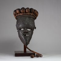 : Masque rituel masculin de haut rang initiatique Salampasu, RD Congo. Le visage est recouvert de feuilles métalliques (cuivre ou laiton). La bouche triangulaire dentée, caractéristique des masques Salampasu, confère à l’objet une forte expressivité rituelle. La coiffe est composée de 28 boules de rotin tressé et la barbe végétale tressée se termine par deux boules identiques. L’intérieur présente