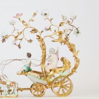 Centre de table à décor de carrosse tiré par quatre chevaux, en bronze ciselé et doré, la scène animée de figures en porcelaine polychrome, représentant deux jeunes enfants dans une calèche ornée de volutes feuillagées et de guirlandes fleuries. Les chevaux sont en porcelaine, fixés sur des bases en porcelaine émaillée à décor fleuri. Travail de style rococo, probablement allemand ou viennois, ver