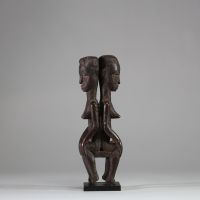 Rare statue Janiforme Akan ( Attié ou Agni) ,Côte d’Ivoire. Bois. Représentation de deux figures féminines assises dos à dos. Les deux têtes présentent une coiffe lobée striée, et les visages sont peints avec des yeux blancs à pupilles noires dilatées. La posture est frontale, hiératique, les mains reposant sur les genoux. La patine est brune à noire, grasse, crouteuse et ancienne, avec traces d'u
