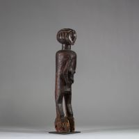 : Ancienne statue féminine Lwalwa, RD Congo. Bois, raphia. Le visage, de forme triangulaire inversée, nez rectiligne et saillant qui remonte jusqu’à la racine des cheveux. Yeux incisés,  bouche discrète et menton légèrement projeté. Le tronc, massif, est flanqué de deux bras longs et cylindriques, plaqués contre les flancs. Les seins sont petits et placés très bas, détail inhabituel qui renforce l