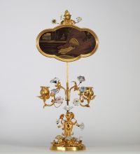 Bougeoir à écran, en bronze ciselé et doré, à décor ajouré de rinceaux feuillagés ponctués de fleurs en porcelaine polychrome de style Meissen. L’ensemble est surmonté d’un écran en laque du Japon de forme chantournée, à décor de paysage lacustre doré dans le goût des exportations de l’époque Meiji. Travail pouvant être attribué à la prestigieuse maison parisienne de l’Escalier de Cristal, vers 18
