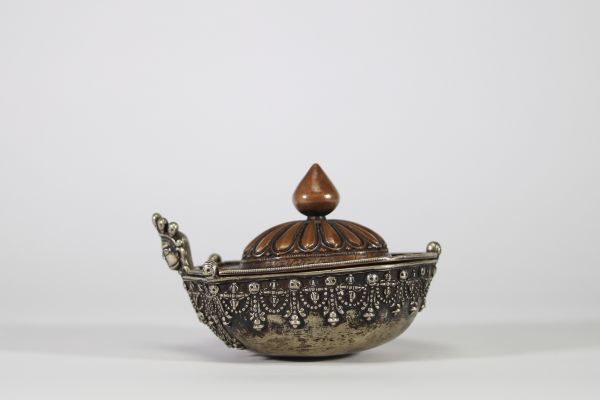 Kapala rituel, Tibet, XVIIIᵉ–XIXᵉ siècle Récipient rituel tibétain de type kapala, composé d’un corps en argent richement décoré de frises de têtes de kirtimukha, de chaînes perlées et de motifs stylisés. Le couvercle est en cuivre martelé, à décor gravé de pétales de lotus rayonnants, surmonté d’un prend finial en forme de flamme, symbole de sagesse dans le bouddhisme tantrique. Aux extrémités, d