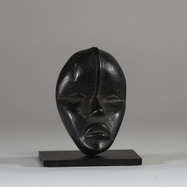Masque passeport Dan, Côte d’Ivoire. Bois. Il présente une esthétique d’une grande pureté. Le visage, de forme ovale, est sculpté avec sobriété : les yeux sont mi-clos et incisés en amande, le nez est droit, triangulaire et structuré, et la bouche légèrement ouverte laisse deviner une expression intériorisée. Une ligne verticale en relief s’élève du haut du nez jusqu’au front, accentuant l’axe de