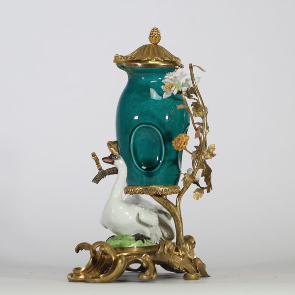 Fontaine à parfum, composée d’un vase en porcelaine de Chine émaillée bleu turquoise, datable du XVIIIe siècle, surmontée d’un couvercle orné d’une pomme de pin dorée. L’ensemble repose sur une base rocaille en bronze ciselé et doré, richement décorée de deux cygnes en porcelaine de Meissen, posés sur des tertres verts. Le décor est complété de fleurs en porcelaine polychrome, probablement de styl