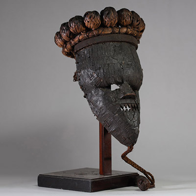 : Masque rituel masculin de haut rang initiatique Salampasu, RD Congo. Le visage est recouvert de feuilles métalliques (cuivre ou laiton). La bouche triangulaire dentée, caractéristique des masques Salampasu, confère à l’objet une forte expressivité rituelle. La coiffe est composée de 28 boules de rotin tressé et la barbe végétale tressée se termine par deux boules identiques. L’intérieur présente