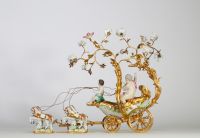 Centre de table à décor de carrosse tiré par quatre chevaux, en bronze ciselé et doré, la scène animée de figures en porcelaine polychrome, représentant deux jeunes enfants dans une calèche ornée de volutes feuillagées et de guirlandes fleuries. Les chevaux sont en porcelaine, fixés sur des bases en porcelaine émaillée à décor fleuri. Travail de style rococo, probablement allemand ou viennois, ver