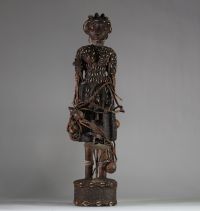 Intéressante statue féminine de type Bocio, Fon, Bénin. Le visage est sculpté de manière stylisée avec un nez proéminent, des yeux en amande profondément incisés, et des lèvres pleines. Une coiffe structurée orne la tête, enrichie de cauris incrustés et de perles. Le cou porte plusieurs rangs de colliers perlés. Le torse et le haut du corps sont entièrement recouverts de cauris patinés, suggérant