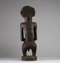 Statue Hemba Singiti, RD Congo. La statue représente une figure masculine debout, dans une posture frontale hiératique. La tête, surdimensionnée, est marquée par une barbe trapézoïdale finement striée, un visage calme aux yeux mi-clos, un nez droit et une bouche discrète. À l’arrière du crâne, une coiffe cruciforme profondément sculptée suggère une coiffure cérémonielle typique. Les bras sont plié
