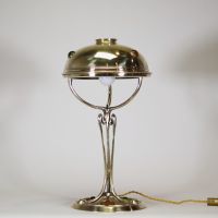 France, vers 1900 Lampe de table Art Nouveau en laiton, capeline ajourée et piétement à cabochons de verre coloré.  Cette lampe décorative de la période Art Nouveau présente une élégante silhouette élancée en laiton doré, avec un fût nervuré à lignes fluides convergeant vers une base circulaire décorée de cabochons de verre sertis en vert, rouge, ambre et jaune, typiques de l’ornementation polychr