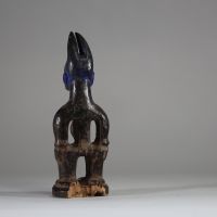 : Statuette Ibéji féminine, Yoruba, Nigéria. Figure féminine debout, dans une posture frontale rigide, les bras éloignés du corps reposant sur des hanches stylisées. Le visage est sculpté avec des yeux en amande, un front bombé, des scarifications linéaires, et une bouche aux lèvres striées horizontalement. Haute coiffe bifide . Patine noire ancienne avec des zones d’usure naturelle aux points de