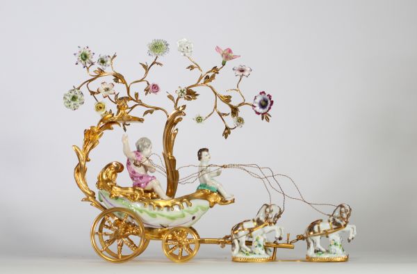 Centre de table à décor de carrosse tiré par quatre chevaux, en bronze ciselé et doré, la scène animée de figures en porcelaine polychrome, représentant deux jeunes enfants dans une calèche ornée de volutes feuillagées et de guirlandes fleuries. Les chevaux sont en porcelaine, fixés sur des bases en porcelaine émaillée à décor fleuri. Travail de style rococo, probablement allemand ou viennois, ver
