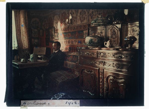 Léon Herlant (1873-1968) — Rare autochrome sur plaque Lumière réalisé en 1912, signé « Herlant », 12 x 9 cm.  Ce précieux portrait montre le photographe lui-même, saisi dans l’intimité de son intérieur raffiné. La scène est magnifiquement composée : les murs sont ornés de tapis kilim aux motifs chatoyants, évoquant un goût prononcé pour l'Orientalisme. Sur des étagères et consoles, s'exposent de d