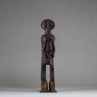 : Ancienne statue féminine Lwalwa, RD Congo. Bois, raphia. Le visage, de forme triangulaire inversée, nez rectiligne et saillant qui remonte jusqu’à la racine des cheveux. Yeux incisés,  bouche discrète et menton légèrement projeté. Le tronc, massif, est flanqué de deux bras longs et cylindriques, plaqués contre les flancs. Les seins sont petits et placés très bas, détail inhabituel qui renforce l