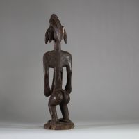 Statue rituelle Bamana « Jonyeleni » , Mali. Sculptée dans un bois ancien à patine brune, représentant une figure verticale aux traits stylisés : coiffe sagittale striée, nez droit, seins coniques scarifiés, ombilic et sexe proéminents. Les mains sont tendues le long du corps avec les paumes ouvertes, dans une posture typique des figures d’enseignement initiatique du Jon.La sculpture présente une
