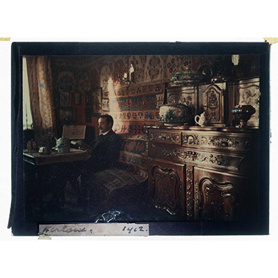 Léon Herlant (1873-1968) — Rare autochrome sur plaque Lumière réalisé en 1912, signé « Herlant », 12 x 9 cm.  Ce précieux portrait montre le photographe lui-même, saisi dans l’intimité de son intérieur raffiné. La scène est magnifiquement composée : les murs sont ornés de tapis kilim aux motifs chatoyants, évoquant un goût prononcé pour l'Orientalisme. Sur des étagères et consoles, s'exposent de d