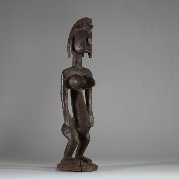 Statue rituelle Bamana « Jonyeleni » , Mali. Sculptée dans un bois ancien à patine brune, représentant une figure verticale aux traits stylisés : coiffe sagittale striée, nez droit, seins coniques scarifiés, ombilic et sexe proéminents. Les mains sont tendues le long du corps avec les paumes ouvertes, dans une posture typique des figures d’enseignement initiatique du Jon.La sculpture présente une