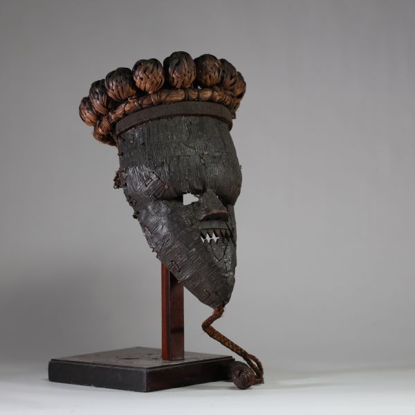 : Masque rituel masculin de haut rang initiatique Salampasu, RD Congo. Le visage est recouvert de feuilles métalliques (cuivre ou laiton). La bouche triangulaire dentée, caractéristique des masques Salampasu, confère à l’objet une forte expressivité rituelle. La coiffe est composée de 28 boules de rotin tressé et la barbe végétale tressée se termine par deux boules identiques. L’intérieur présente