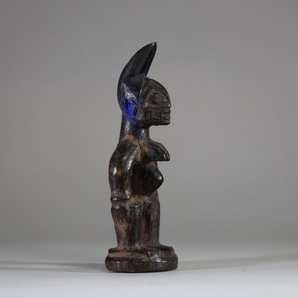 : Statuette Ibéji féminine, Yoruba, Nigéria. Figure féminine debout, dans une posture frontale rigide, les bras éloignés du corps reposant sur des hanches stylisées. Le visage est sculpté avec des yeux en amande, un front bombé, des scarifications linéaires, et une bouche aux lèvres striées horizontalement. Haute coiffe bifide . Patine noire ancienne avec des zones d’usure naturelle aux points de