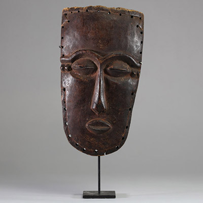 Masque facial anthropomorphe Lele ( bashilele), RD Congo, région du Kasaï. Bois présentant une surface patinée ancienne, plus brillante et lisse dans les zones exposées. Trous sur les bords permettant l’attache d’ornements ou a une structure. Yeux sculptés en amande avec fente étroite. La bouche est petite et sculptée en légère saillie. Le nez est droit et allongé avec une base marquée. Exemplaire
