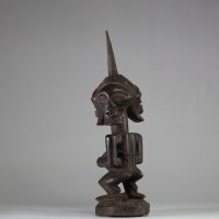 Puissant fétiche janiforme Songye, Congo RDC. Représentant une figure accroupie, aux bras anguleux et au torse tendu, coiffée d’une crête sagittale striée proéminente. La tête présente trois visages distincts disposés en triangle, chacun aux yeux mi-clos et à la bouche souriante, exprimant la vigilance et la vision multiple. Ce type de figure janiforme tripartite, rare dans l’art songye, renvoie à