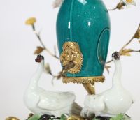 Fontaine à parfum, composée d’un vase en porcelaine de Chine émaillée bleu turquoise, datable du XVIIIe siècle, surmontée d’un couvercle orné d’une pomme de pin dorée. L’ensemble repose sur une base rocaille en bronze ciselé et doré, richement décorée de deux cygnes en porcelaine de Meissen, posés sur des tertres verts. Le décor est complété de fleurs en porcelaine polychrome, probablement de styl