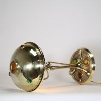 France, vers 1900 Lampe de table Art Nouveau en laiton, capeline ajourée et piétement à cabochons de verre coloré.  Cette lampe décorative de la période Art Nouveau présente une élégante silhouette élancée en laiton doré, avec un fût nervuré à lignes fluides convergeant vers une base circulaire décorée de cabochons de verre sertis en vert, rouge, ambre et jaune, typiques de l’ornementation polychr