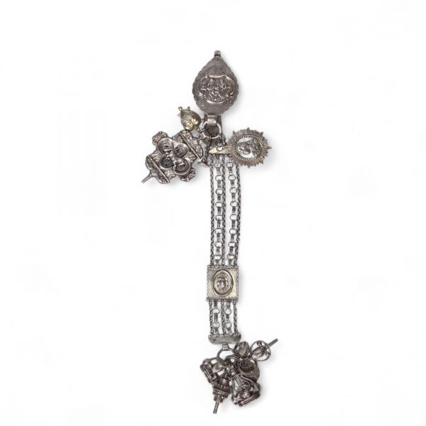 Chatelaine en argent, Pays-Bas, XIXᵉ siècle Chatelaine en argent composée de plusieurs éléments richement décorés, réunis par une double chaîne à maillons ciselés. L’ensemble est orné de médaillons à motifs en bas-relief, de pendentifs fonctionnels (grelots, sifflet, étui, petite théière ou vinaigrier...), et d’un fermoir principal gravé de scènes figuratives.  Le poinçon au lion marchant n°2, vis