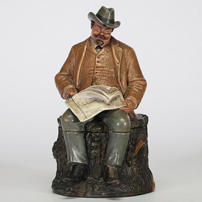 Bernard Bloch (atelier BB), Bohême, fin XIXe – début XXe siècle Pot à tabac en terre cuite polychrome figurant un homme assoupi vêtu d’un costume campagnard, assis sur une souche, les yeux mi-clos, un journal déployé sur les genoux.