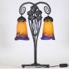 Muller Frères, Lunéville, vers 1905 Lampe à deux lumières en fer forgé martelé à décor de ginkgo biloba et verreries multicouches signées.  Superbe lampe Art Nouveau en fer forgé patiné noir, montée sur une base rectangulaire, s’élevant en double col de cygne à volutes stylisées. Le fût est orné de feuilles et hampes florales de ginkgo biloba, motif emblématique du symbolisme végétal de la Belle É