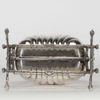 Élégante boîte à biscuits en métal argenté, à ouverture double, dite « shell biscuit box », prenant la forme d’un coquillage stylisé monté sur un piètement ajouré à motifs de branches naturalistes croisées. Le couvercle à charnières s’ouvre de part et d’autre pour dévoiler deux compartiments intérieurs à décor ajouré d’entrelacs floraux et géométriques. L’ensemble repose sur quatre pieds en forme