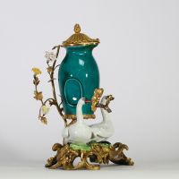 Fontaine à parfum, composée d’un vase en porcelaine de Chine émaillée bleu turquoise, datable du XVIIIe siècle, surmontée d’un couvercle orné d’une pomme de pin dorée. L’ensemble repose sur une base rocaille en bronze ciselé et doré, richement décorée de deux cygnes en porcelaine de Meissen, posés sur des tertres verts. Le décor est complété de fleurs en porcelaine polychrome, probablement de styl