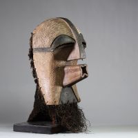 Très beau masque Songye Kifwebe avec sa coiffe d’origine . Rd Congo. Bois et fibres. Morphologie typique des masques Kifwebe : visage allongé avec striures régulières en chevrons couvrant l’ensemble de la surface faciale, nez proéminent en arête verticale, yeux mi-clos formant des fentes horizontales sombres, bouche tubulaire sculptée en forme d’étoile percée. La partie inférieure triangulaire est