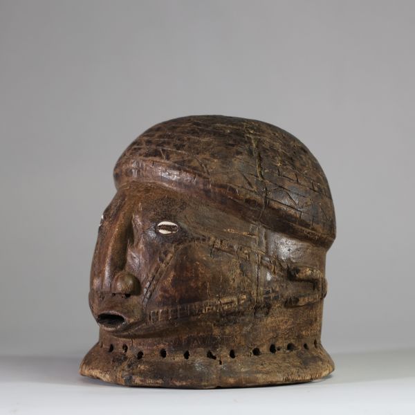 Masque-casque en bois, de forme hémisphérique Tabwa, RD congo. Visage sculpté avec un nez droit, saillant, de forme anguleuse, très caractéristique des styles Tabwa et Luba-Tabwa. Les yeux sont incrustés de cauris, Les scarifications faciales linéaires et parallèles, courant des tempes vers la bouche, sont typiques des traditions Tabwa. La patine naturelle, les abrasions autour de la base perforée