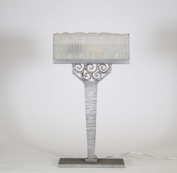 France, vers 1930 Lampe de bureau Art Déco en métal martelé nickelé et verre pressé à décor floral et géométrique.  Reposant sur une base rectangulaire, cette lampe présente un fût élancé à surface martelée, d’une sobriété élégante, souligné d’une finition nickelée mate. L’ensemble est rehaussé d’un nœud ornemental de volutes forgées à la main, typique du vocabulaire décoratif de l’époque.  L’abat