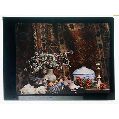 Léon Herlant (1873-1968) — Remarquable autochrome sur plaque Lumière, 8,5 x 10 cm.  Cette somptueuse nature morte s'inscrit dans la tradition des peintures flamandes du XVIIᵉ siècle par la richesse de sa composition et la subtilité de sa lumière. Au centre, un élégant candélabre en argent capte la lumière naturelle et l’irradie délicatement sur la scène, révélant avec finesse les détails des ailes