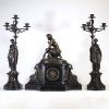 Garniture de cheminée en bronze patiné et marbre noir, signée Delvaulx, époque Napoléon III Imposante garniture de cheminée en bronze patiné et marbre noir composée d’une pendule centrale surmontée d’une figure féminine nue, assise dans une posture de toilette, finement sculptée et reposant sur un socle tripartite orné de bas-reliefs à sujet antique. Elle est accompagnée d’une paire de candélabres